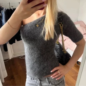 Grå offshoulder stickad topp - Mysig grå stickad topp med offshoulder-design och kort ärm. Toppen har en fluffig, mjuk yta och sitter tight mot kroppen. Perfekt för dig som vill ha en trendig och bekväm look.
