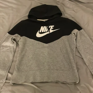 Nike hoodie i svart och grå - Nike hoodie med svart ovandel och grå nederdel. Stor vit Nike-logga på bröstet, huva med snörning och känguruficka framtill. Perfekt för chill eller träning.