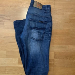 Nudie Jeans blåa Thin Finn  - Säljer ett par Nudie Jeans Thin Finn i mörkblå tvätt med klassisk femficksdesign. Jeansen är tillverkade i stretchig denim av 98,5% ekologisk bomull och 1,5% elastan. Snygga slitningar och detaljer, perfekt för dig som gillar en smalare passform. Dom är i storlek 29/32 och är sönder i dragkedjan(se sista bilden)