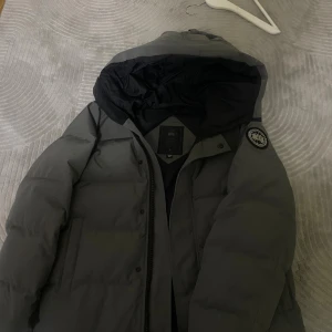 Grå dunjacka från Canada Goose - Säljer en grå dunjacka från Canada Goose med svart innerfoder och huva. Jackan har klassisk patch på ärmen, dragkedja framtill och en clean, stilren look. Perfekt för kalla dagar och riktigt snygg till streetwear. Använd förra vintern, macmillan.