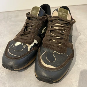 Valentino Garavani Rockrunner sneakers - Snygga Valentino Garavani Rockrunner sneakers med camouflagemönster i brunt, beige, svart och grått. Skorna har detaljer i mocka och skinn, snörning och platt sula. Röd detalj på hälen ger extra edge. Perfekta för dig som gillar exklusiva streetwear-vibes. Liten defekt på insidan av höger sko men inget som stör väldigt mycket. Innermått 27,3 cm 