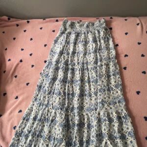 Blåvit långkjol från 157, XS - Långkjol från 157 i storlek XS med blått och vitt blommigt mönster. Kjolen har smock i midjan för en bekväm passform och är tillverkad i ett lätt och luftigt tyg, perfekt för varma dagar. Den har en flowy och avslappnad siluett.