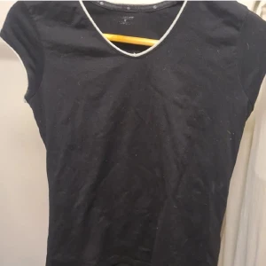 Svart topp - Svart topp med vit kant längs v-ringningen och ärmsluten. Toppen är ärmlös och har en enkel. Perfekt för en clean och avslappnad look.