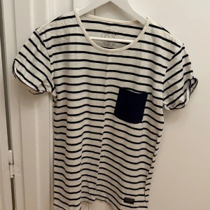 Randig t-shirt från Cottonfield - Snygg vit t-shirt med marinblå ränder från Cottonfield. Klassisk rund halsringning, korta ärmar och en marinblå bröstficka som extra detalj. Tillverkad i 100% bomull för en skön och luftig känsla. Perfekt för en avslappnad och stilren look. Storlek M.