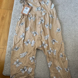 Beige jumpsuit med fjärilar från H&M - Superfin beige jumpsuit från H&M i storlek 92, täckt med vita och svarta fjärilar. Gjord i mjuk bomull och har knappar framtill. Perfekt för dig som gillar lekfulla och bekväma plagg med snyggt mönster.