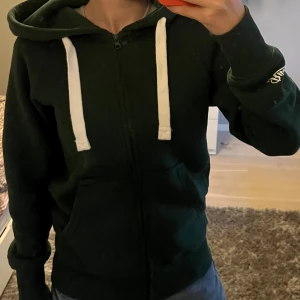 Mörkgrön hoodie från Russemerch - Mörkgrön hoodie från Russemerch med vita snören, dragkedja och huva. Tröjan har fickor framtill och logga broderad på ärmen. Perfekt för chill dagar och passar till jeans eller mjukisbyxor.
