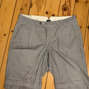 Randiga shorts från Blueprint, M - Snygga randiga shorts från Blueprint i grått och vitt. Klassisk modell med fickor fram och bak, bälteshällor och knappstängning. Tillverkade i 100% bomull för en skön känsla. Perfekta för varma dagar när du vill ha en clean och stilren look. Storlek M.