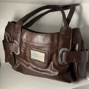 Brun handväska från D&G i skinn - Snygg brun handväska från D&G i mjukt skinn med metallplatta framtill och coola detaljer på sidorna. Väskan har två rejäla handtag och en stilren, klassisk form som passar till många outfits. Perfekt för dig som gillar designer och vill sticka ut med vintage stilen.