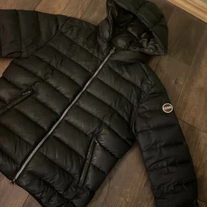 Svart dunjacka med huva från Colmar - Svart dunjacka från Colmar med huva och två dragkedjeförsedda fickor. Jackan har ett stilrent, quiltat mönster och logotyp på ärmen. Perfekt för kalla dagar och har en snygg, klassisk look som funkar till det mesta. Några smutsfläckar kan också kanske vara defekter(kolla två sista bilderna) men om det endast är smuts så går det bort. Storlek 44 som motsvarar ca XS/S