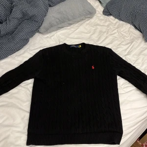 Svart stickad tröja från Ralph Lauren - Säljer en klassisk svart stickad tröja från Ralph Lauren. Den har ett diskret kabelstickat mönster och den ikoniska röda loggan på bröstet. Perfekt för både höst och vinter, med ribbade muddar och nederkant för extra komfort. Passar lika bra till vardags som till lite finare tillfällen.