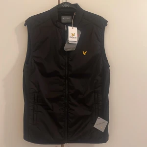 Svart dunväst från Lyle & Scott - Svart dunväst från Lyle & Scott med gul logga på bröstet. Västen har hög krage, dragkedja framtill och två fickor på sidorna. Tillverkad i syntetmaterial och har en clean, sportig look som passar perfekt till lager-på-lager-stil.