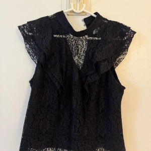 Svart spetsblus med volangdetaljer - Supersnygg svart blus i spets med volangdetaljer längs axlar och framstycke. Blusen är ärmlös och har hög hals med knäppning bak. Perfekt för dig som gillar romantisk och trendig stil. Spetsen har ett vackert blommönster som ger en lyxig känsla. Passar typ S-M