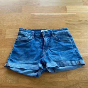 Blå denimshorts med upprullad kant - Blå denimshorts med upprullad kant nertill. Shortsen har bälteshällor, knapp- och dragkedjestängning samt raka ben. 