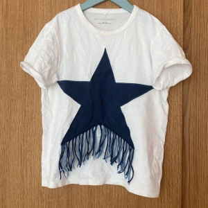 Vit t-shirt med blå stjärna Stella McCartney - Vit t-shirt från Stella McCartney med en stor mörkblå stjärna framtill. Stjärnan har coola fransar som hänger ner. T-shirten är kortärmad och i mjuk bomull, perfekt för en avslappnad och trendig look. Original pris 500. Kan ksk förhandla om pris