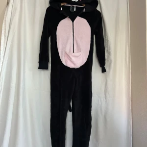 Svart katt-onesie från H&M med huva - Mysig svart onesie från H&M med rosa mage, broderat kattansikte på huvan, öron och löstagbar svans. Dragkedja framtill och långa ärmar med muddar. Perfekt för chilliga dagar eller temakvällar.