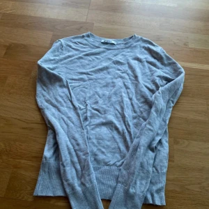 Ljusgrå stickad tröja med rund hals - Ljusgrå stickad tröja med rund halsringning och långa ärmar. Tröjan har breda ribbade muddar vid ärmslut och nederkant. 