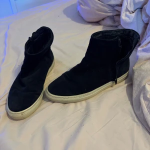 Svarta mockaboots från Filippa K - Svarta boots från Filippa K i mjuk mocka med vit platt sula. Skorna har dragkedja på sidan för enkel påtagning och en minimalistisk design. Insidan är fodrad för extra komfort. Perfekta för dig som gillar stilrena och klassiska skor. Dom är i storlek 41 men passar mer som stor 39 eller vanlig 40