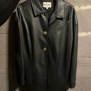 Svart skinnkappa - Tunn trench coat liknande faux skinn jacka från NA-KD. Tillhörande skärp tillkommer. Jackan är knälång ungefär. 