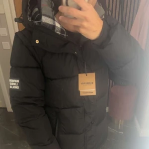 Svart jacka/dunväst från Burberry - Svart dunväst från Burberry med klassiskt rutigt foder i huvan och guldiga knappar. Västen har hög krage, avtagbara ärmar och en snygg logga på vänster axel. Perfekt för lager-på-lager och streetstyle.