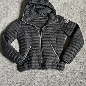 Svart pufferjacka från Colmar storlek xs - Snygg svart pufferjacka från Colmar med huva och två dragkedjeförsedda fickor. Jackan har quiltad design, Colmar-logga på ärmen och hel dragkedja framtill. Perfekt för kyliga dagar och riktigt lätt att bära.