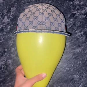 GG keps med GG-mönster och röd-blå rand - Säljer en snygg GG keps i beige med blått GG-monogram över hela kepsen. Kepsen har en klassisk böjd skärm, justerbart band bak i blått och en röd-blå rand på sidan. Tillverkad i bomullscanvas med detaljer i skinn.