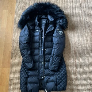 Svart lång dunjacka Rockandblue 38 - Snygg svart lång dunjacka från Rockandblue med quiltade detaljer, stor fluffig fuskpäls på huvan och dragkedja framtill. Jackan har en badge på ärmen, flera fickor och är perfekt för kalla dagar. Klassisk och stilren modell med schyssta detaljer.