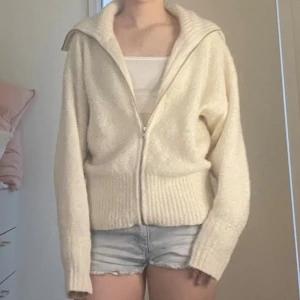 Beige stickad kofta från Esprit - Mysig beige stickad kofta från Esprit med dragkedja framtill och bred krage. Koftan har långa ärmar och ribbade muddar vid ärmslut och nederkant. Perfekt att slänga på sig när det är lite kyligt ute.