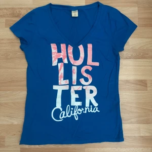 Blå Hollister t-shirt med tryck - Blå t-shirt från Hollister med stort tryck i rosa och vitt framtill där det står 'Hollister California'. T-shirten har v-ringning och korta ärmar. Materialet är mjuk bomull och trycket har palmmotiv i bokstäverna. Använd en gång.