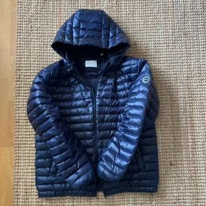 Marinblå dunjacka från GANT med huva - Snygg marinblå dunjacka från GANT med huva och dragkedja framtill. Jackan har quiltad design, rund GANT-logga på ärmen och är lätt vadderad. Perfekt för kyliga dagar och enkel att matcha med olika outfits.