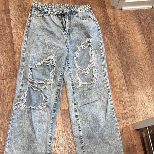 Ljusblå slitna jeans med raka ben - Säljer ett par ljusblå jeans med kraftiga slitningar och hål på båda benen. Jeansen har raka ben, klassisk femficksmodell och hög midja. Perfekta för en avslappnad och trendig streetstil.