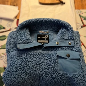 Blå fleece från Pelle P med ficka storlek 150 - Mysig blå fleece från Pelle P i storlek 150 med hög krage och tryckknappar. Tröjan har en bröstficka med lock och knapp, samt ett fluffigt teddy-material som håller dig varm. Perfekt för kyliga dagar och enkel att dra på sig över en t-shirt eller en tunnare långärmade tröja.