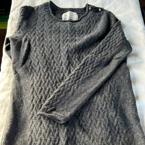 Grå stickad tröja från Newhouse - Mysig grå stickad tröja från Newhouse med flätstickat mönster och knappar på axeln. Tröjan är tillverkad i 80% lammull och har långa ärmar. En liten small så här av dig om du vill ha mått