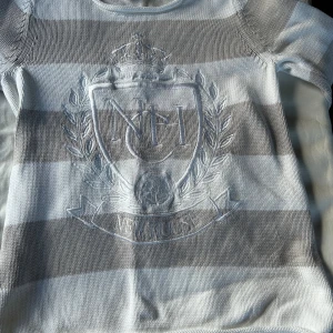 Randig stickad tröja från Newhouse - Snygg stickad tröja från Newhouse i breda grå och vita ränder. Tröjan har broderat emblem framtill, rund halsringning och långa ärmar. Tillverkad i 100% bomull för en skön och mjuk känsla. Perfekt för dig som gillar klassisk och preppy stil.