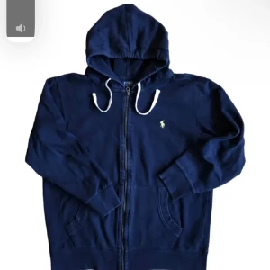 Marinblå hoodie från Polo Ralph Lauren - Snygg marinblå hoodie från Polo Ralph Lauren med dragkedja framtill, vita snören i huvan och klassisk logga broderad på bröstet. Perfekt för en avslappnad stil och enkel att matcha med jeans eller joggers. Står L, men passar som xs