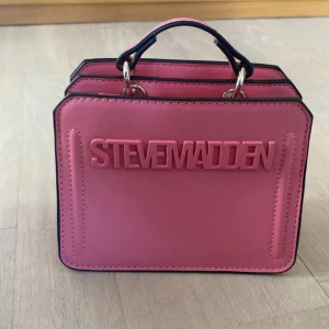 Rosa handväska från Steve Madden -  Helt oanvänd steve madden väska med tydlig logga framtill. Väskan har en bred, justerbar axelrem med mönster, det går att använda väskan både med och utan axelremmen. Väskan har två fack inuti, en med blixtlås och en utan. Väskan har silverfärgade detaljer och magnetlås. Skriv för fler bilder eller vid frågor!