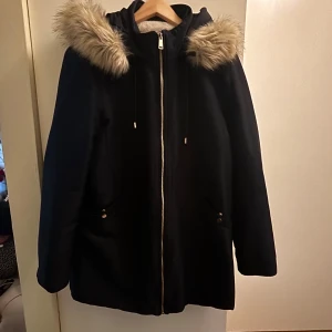 Svart parkajacka med fuskpäls från MNG - Svart parkajacka från MNG med beige fuskpäls på huvan och mjukt teddyfoder på insidan. Jackan har dragkedja framtill, två fickor med knappar och snörning vid huvan. Perfekt för kalla dagar med sin varma och mysiga känsla.