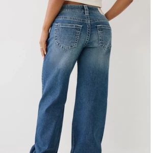 True Religion bobby jeans blå  - Säljer ett par blå jeans från True Religion med ikoniska bakfickor och kontrastsömmar. Jeansen har en bred, loose passform och klassisk mellanblå tvätt med slitningar. Perfekta för dig som gillar streetstyle och vill sticka ut med snygga detaljer. (Dom är lite förkortade) Köpta för 1299kr inne i raglady.
