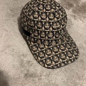 Beige och svart keps Salvatore Ferragamo - Snygg keps från Salvatore Ferragamo i beige och svart med ikoniskt mönster över hela kepsen. Tillverkad i bomull med svart läderdetalj baktill där loggan är ingraverad. Perfekt accessoar för att lyfta din outfit med lyxig känsla. Har ni några frågor eller funderingar tveka inte för att höra av er 😁😁