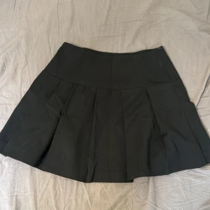 Svart plisserad kortkjol - Snygg svart kortkjol med bred linning och plisserade detaljer som ger extra volym. Kjolen har en klassisk och stilren look, perfekt att matcha med en crop top eller oversized hoodie. Materialet känns mjukt och följsamt.