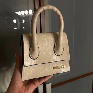Beige handväska från Jacquemus - Superfin beige handväska från Jacquemus med croco-mönstrad yta och guldiga detaljer. Väskan har en stilren, boxig form och två rejäla handtag. Perfekt för dig som vill ha en trendig accessoar med lyxig känsla.