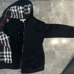 Svart hoodiejacka från Burberry - Svart hoodiejacka från Burberry med klassiskt rutigt foder i huvan. har dragkedja framtill, snörning i huvan och broderad logga på bröstet. Tillverkad i mjuk bomull och polyesterblandning. Perfekt för dig som vill ha en stilren och lyxig look. Väldigt varm nu för vintern 