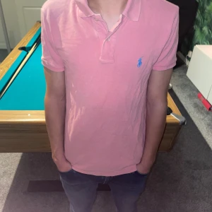 Rosa pikétröja från Ralph Lauren - Säljer en klassisk rosa pikétröja från Ralph Lauren med blå broderad logga på bröstet. Tröjan har korta ärmar, krage och knappar framtill. Perfekt för dig som gillar stilren och tidlös design.