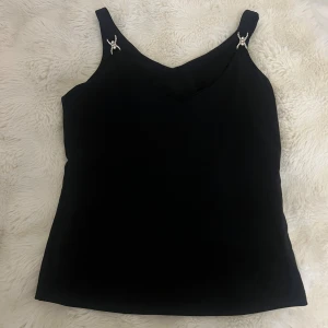 Svart linnetopp med strassdetaljer H&M - Svart linnetopp från H&M med breda axelband och snygga strassdetaljer på axelbanden. Toppen har en enkel, rak passform och v-ringning framtill. Perfekt för dig som vill ha en stilren men ändå glittrig look.