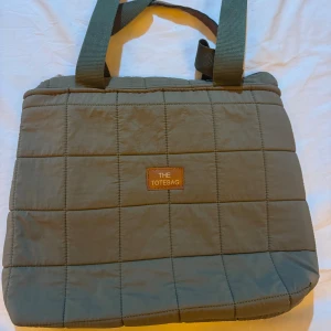 Quiltad grön shoppingväska The Totebag - Stilren quiltad shoppingväska i olivgrönt tyg från The Totebag. Väskan har breda axelband och rutmönster över hela ytan. Perfekt för att bära med sig allt man behöver under dagen.