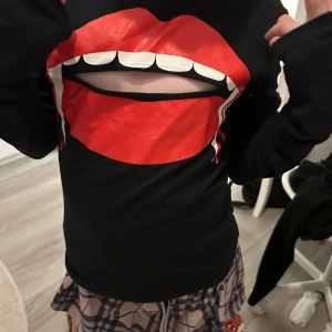 Svart tröja med röd läpp-tryck - Cool svart långärmad tröja med stort tryck av röda läppar och vita tänder på framsidan. Tröjan har en cut-out detalj mitt i munnen som ger en edgy look. Perfekt för dig som vill sticka ut med din stil.