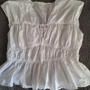 Vit blus med volang från H&M - Säljer en luftig vit blus från H&M i storlek M. Blusen har volang nertill, knytning framtill och är ärmlös med lätt rynkade detaljer. Perfekt för en fräsch och somrig look. Aldrig använd dock har jag tagit bort prislappen men helt oanvänd 