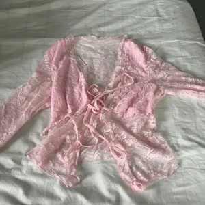 Rosa spetsig bolerotopp med knyt - Säljer en söt och romantisk bolerotopp i ljusrosa spets med blommigt mönster. Toppen har långa ärmar och knyt framtill för en extra feminin touch. Perfekt att styla över ett linne eller klänning för en trendig look.