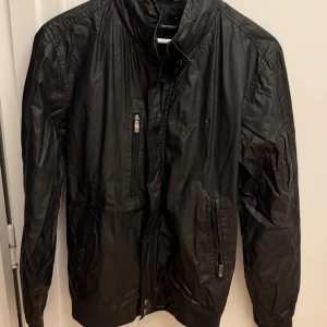 Svart bomberjacka Peak Performance - Svart bomberjacka från Peak Performance med glansig finish, flera dragkedjeförsedda fickor och ribbade muddar. Jackan har hög krage, avtagbar huva och klassisk bomberstil. Perfekt för dig som gillar en sportig och clean look. Storlek M.