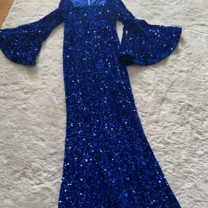 Blå glittrig långklänning med trumpetärm - Säljer en maxad blå långklänning täckt av glittriga paljetter. Klänningen har vida trumpetärmar och markerad midja, med dragkedja i ryggen. Perfekt för dig som vill sticka ut och glänsa! Fodrad upptill för extra komfort.