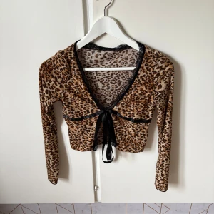 Leopardmönstrad bolerotopp från SHEIN - Säljer en croppad bolerotopp från SHEIN i leopardmönster med lång ärm. Toppen har svart spetskant vid halsen och en snygg knytning framtill med satinband. Materialet är mjukt och stretchigt polyester, perfekt för att ge din outfit lite edge.